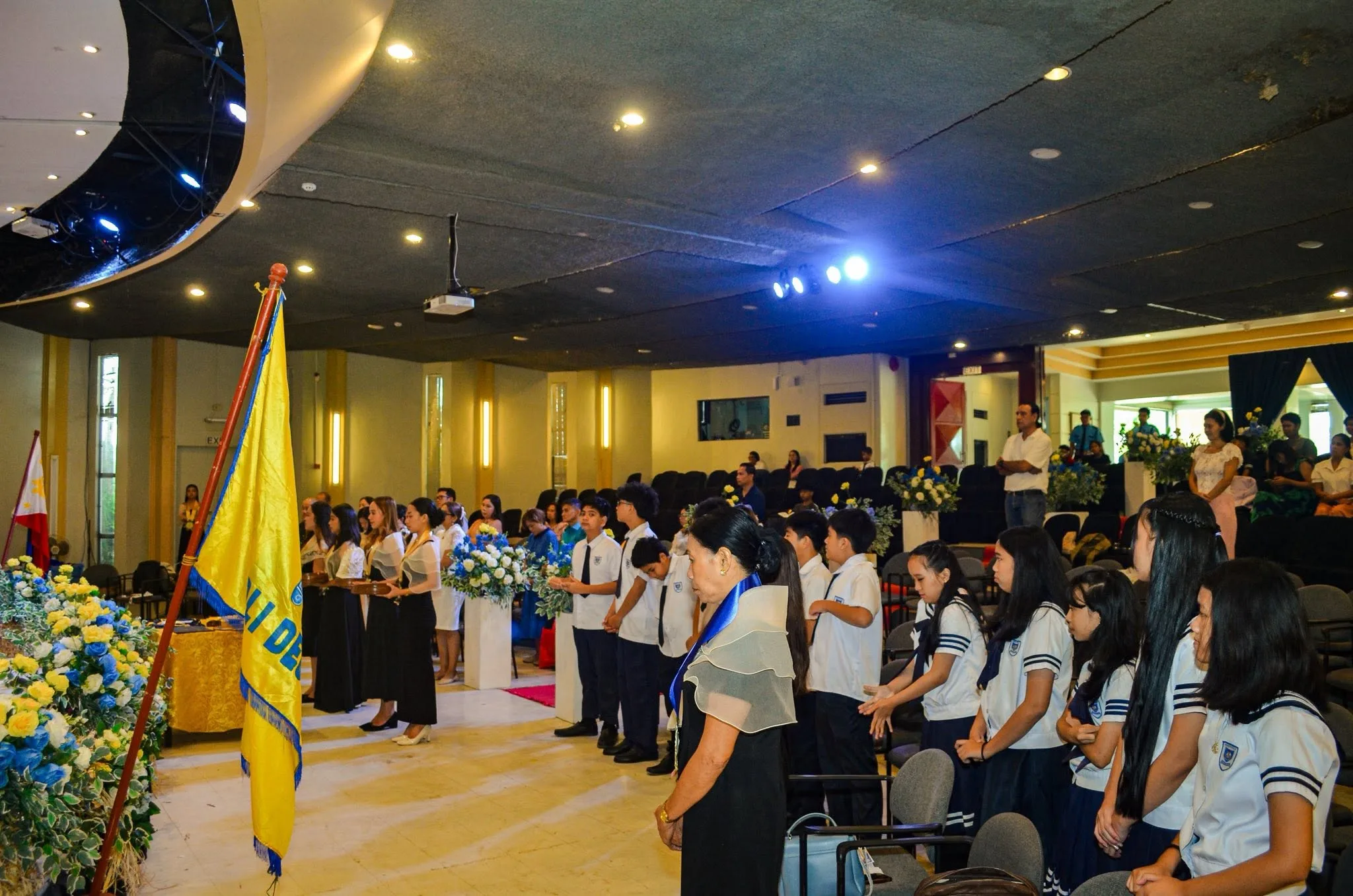 Students in white and navy uniforms stand in rows during a formal school assembly, with the Northville school flag and the Philippine flag at the front of the hall.
