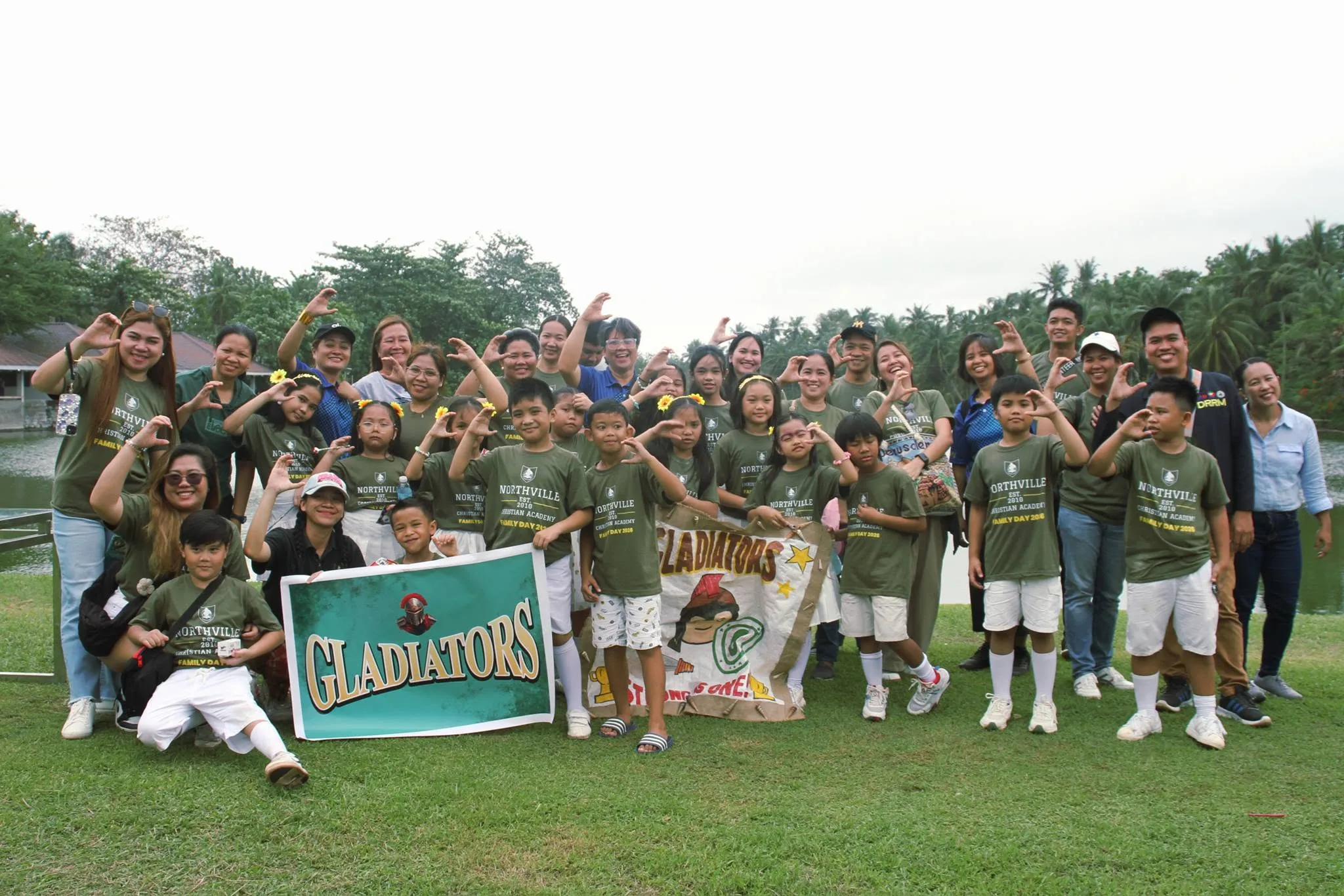 Families, students and teachers in green Northville Family Day shirts crowd together for a group photo behind a Gladiators banner, many making heart shapes with their hands.