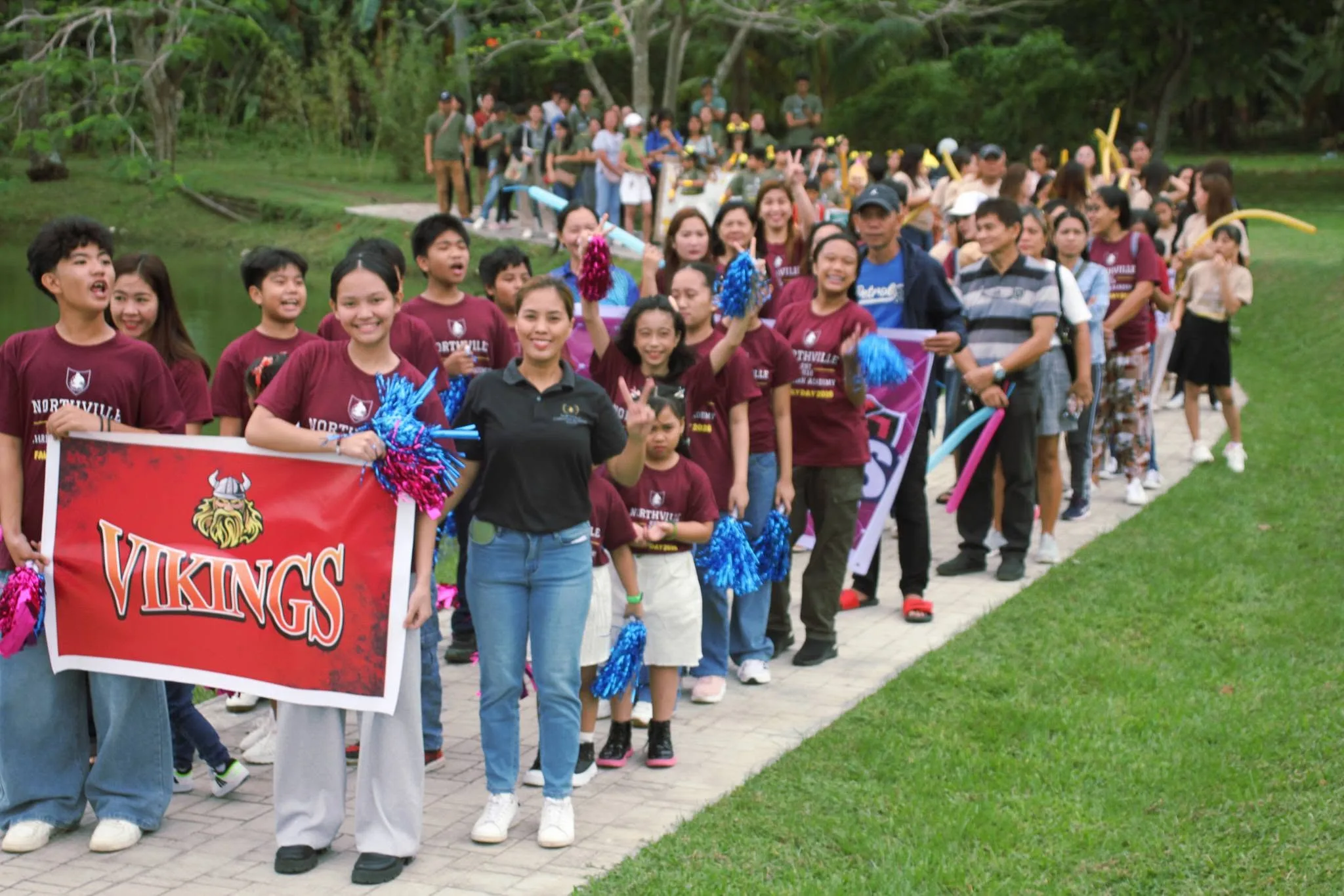Students in maroon Northville shirts march along a lakeside path behind a red Vikings banner, waving pom-poms during the Family Day parade.