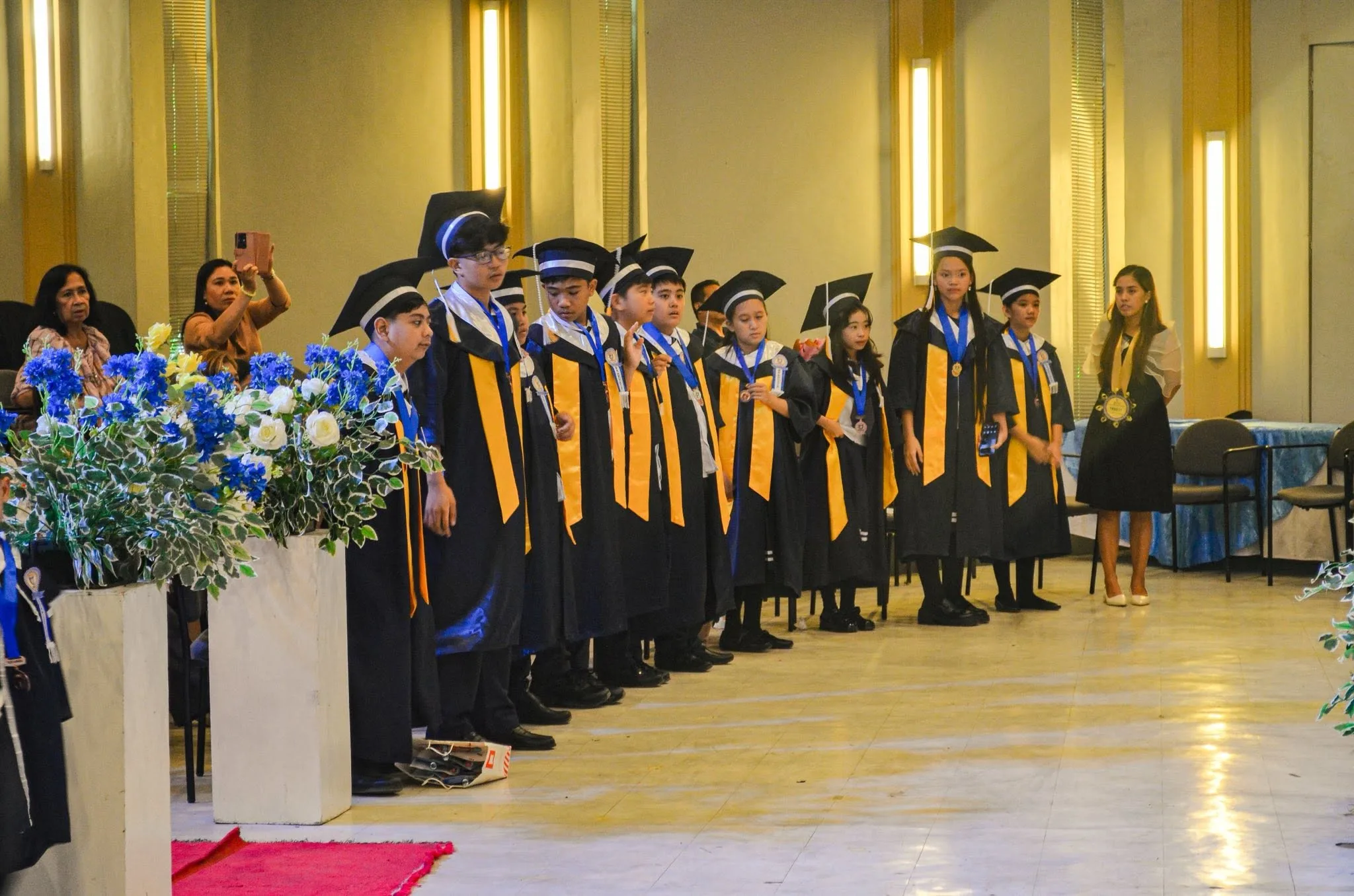 Graduating students in black caps and gowns with gold sashes stand together at their commencement ceremony.