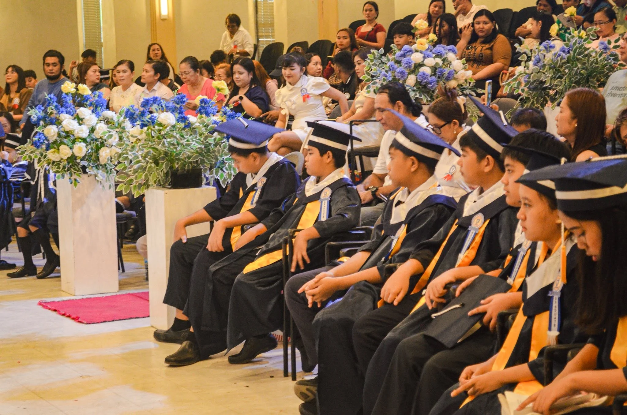 Rows of graduates in caps and gowns stand for a formal graduation ceremony, with flower arrangements on stage.