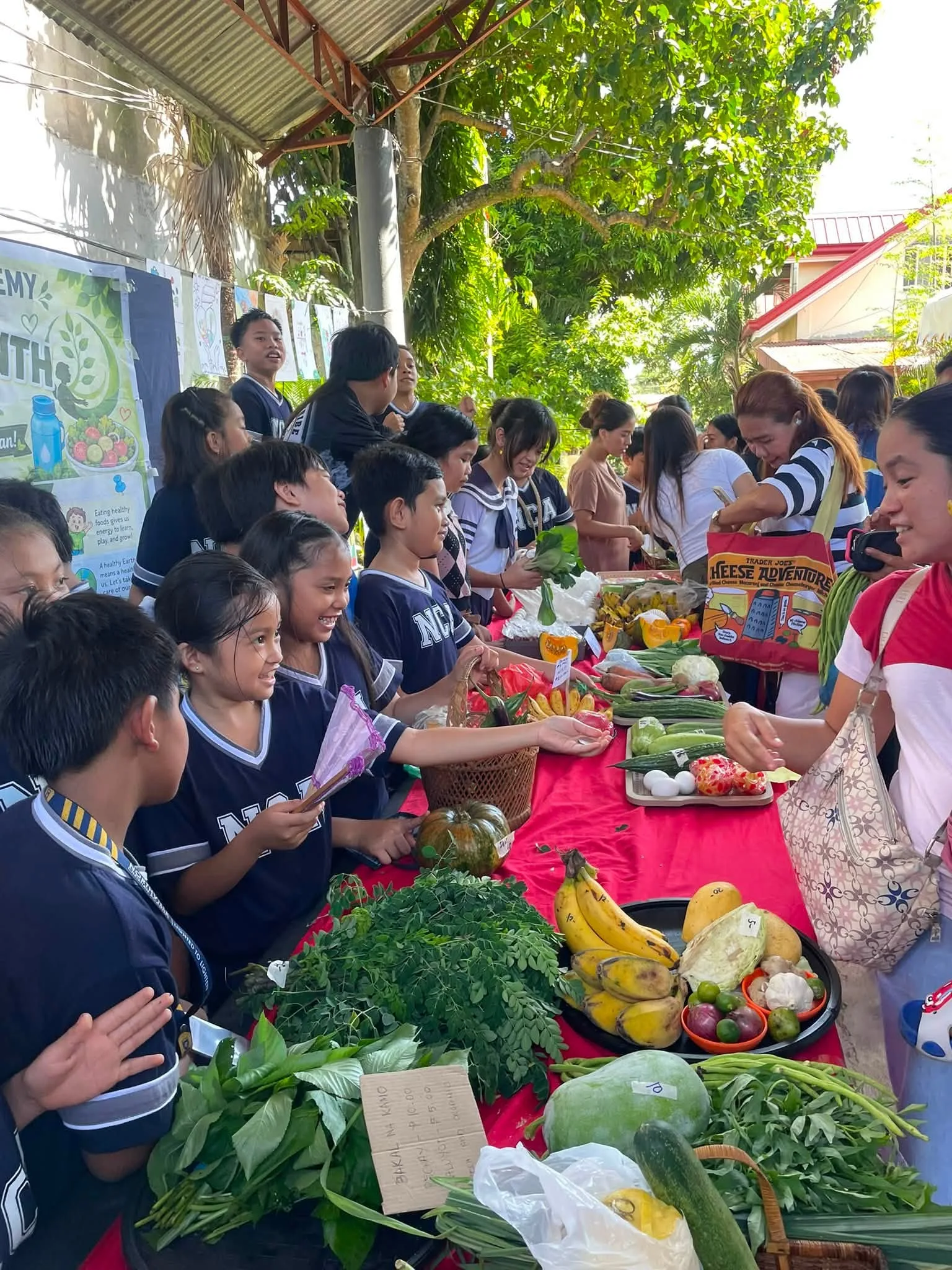 Students in navy uniforms sell vegetables, bananas and greens across a market table to parents during Nutrition Market Day.