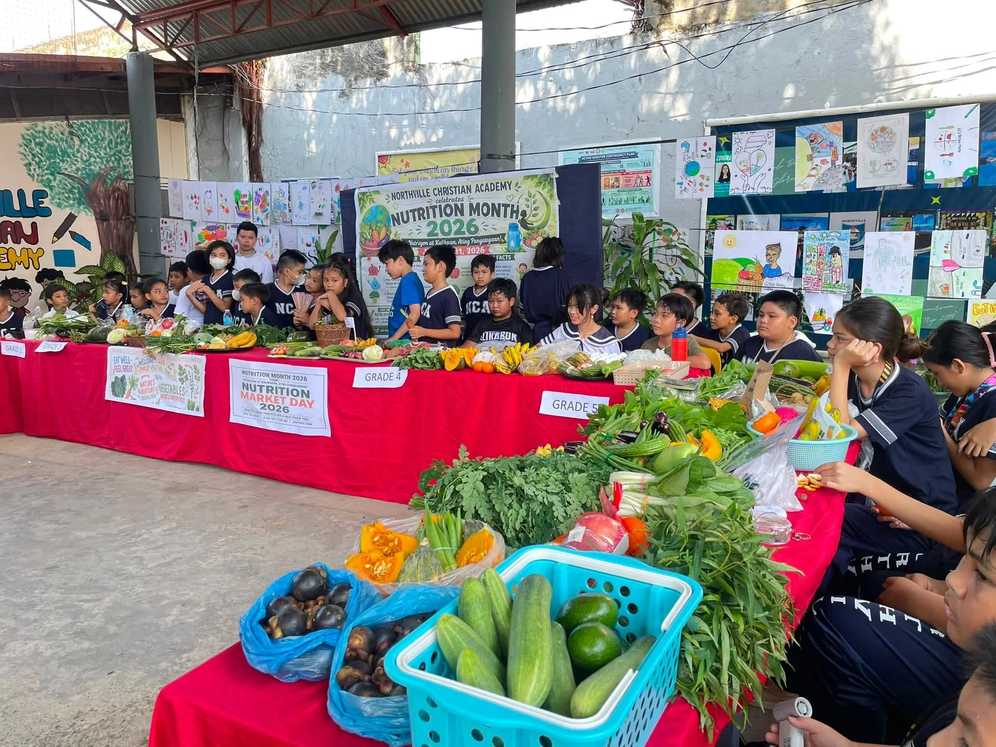 Children gather around a table of fruits and vegetables arranged for a Nutrition Month display, with posters behind them.