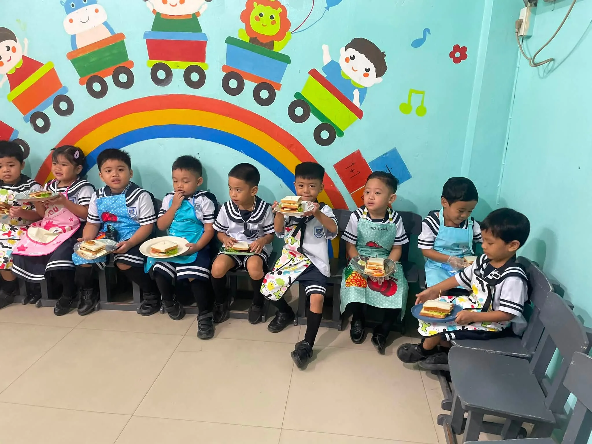 Preschoolers in sailor-collar uniforms and aprons sit along a classroom wall painted with a rainbow and a train, eating sandwiches from paper plates.