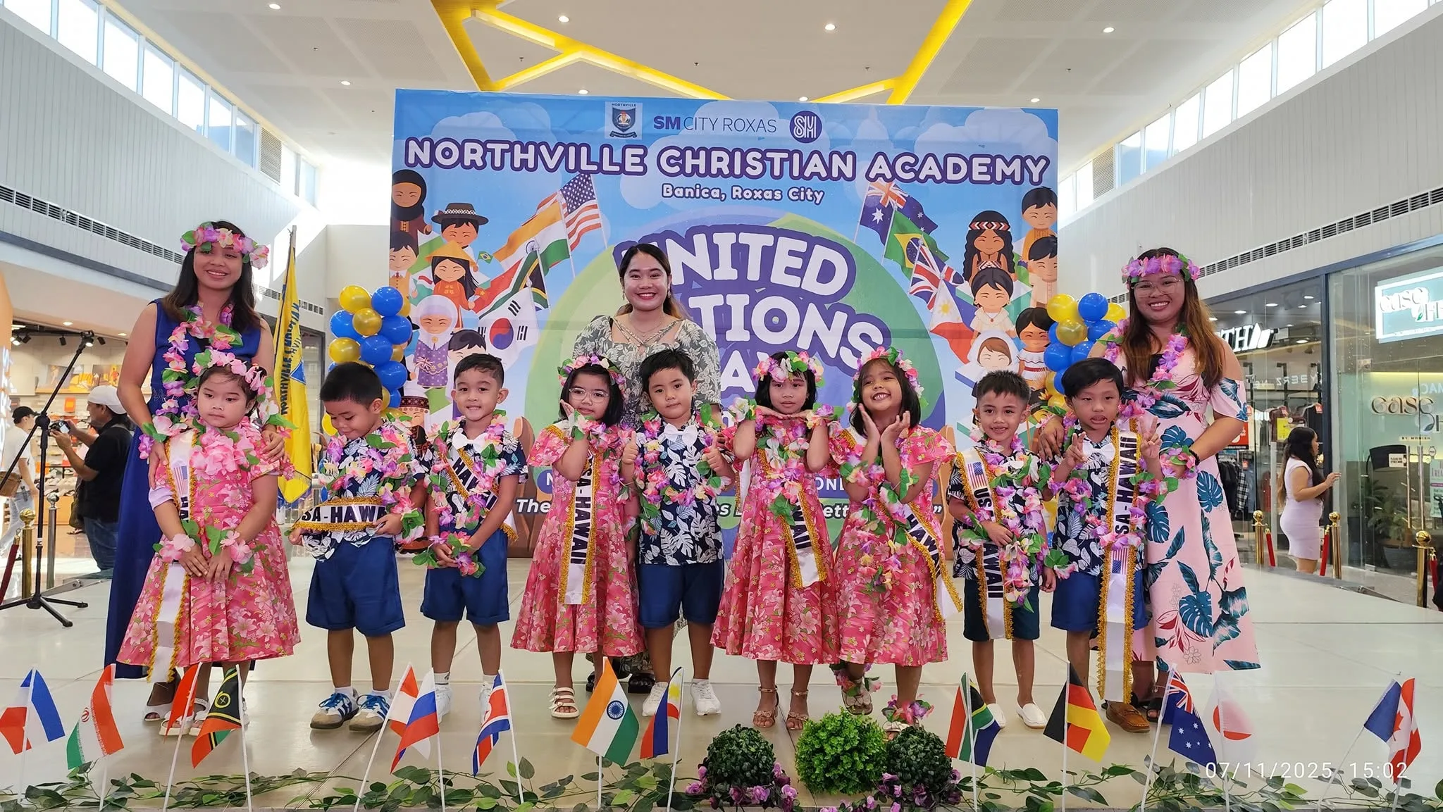 Preschoolers in floral leis and Hawaiian shirts stand with three teachers on a United Nations Day stage at SM City Roxas.