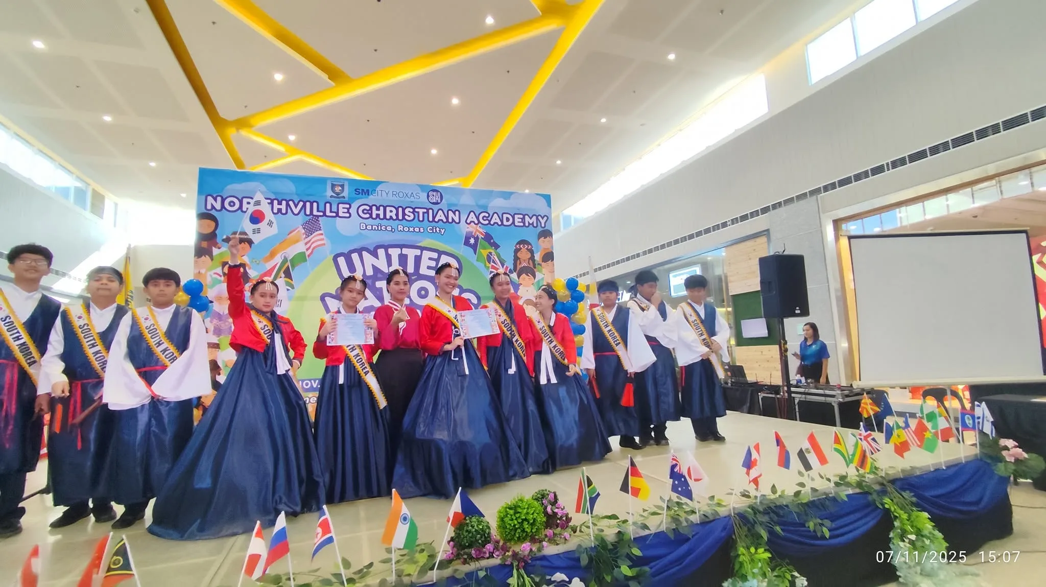 Older students in South Korean hanbok hold certificates on stage during United Nations Day at SM City Roxas.